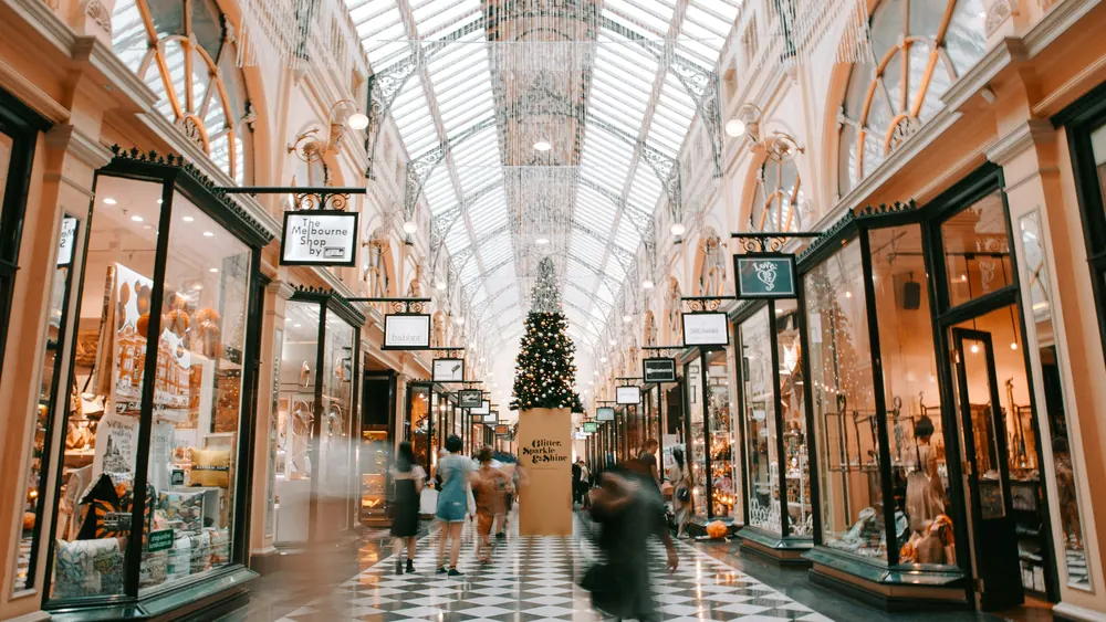 A grand retail arcade under a glass roof, shoppers between lit storefronts